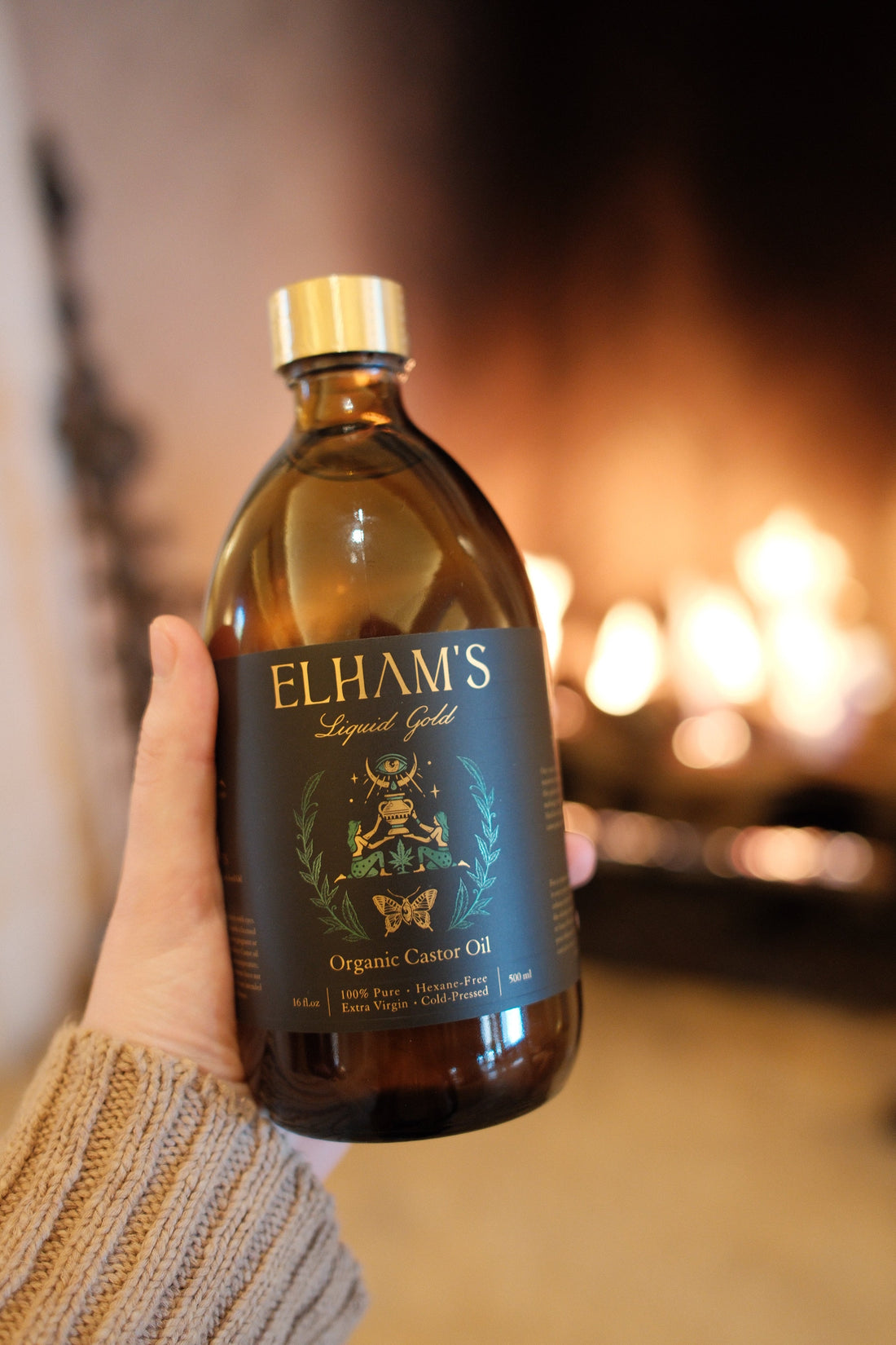 A hand holding a bottle of Elham’s Organic Castor Oil beside a warm, glowing fireplace